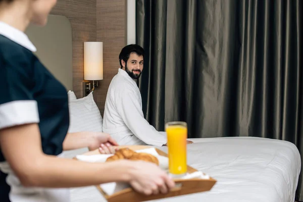 Обрезанный снимок горничной в форме, держащей поднос с круассанами и соком для гостя отеля, пока он сидит на кровати — стоковое фото
