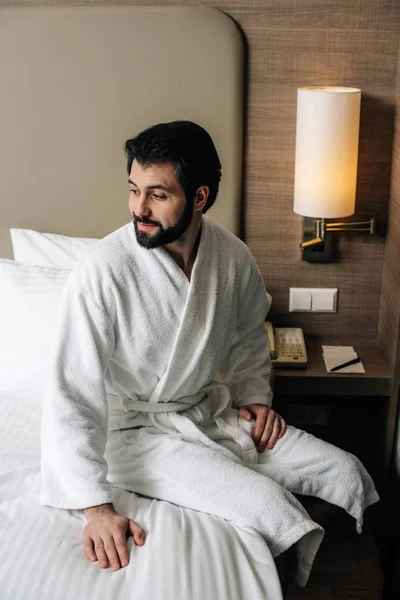 Hombre sonriente en albornoz sentado en la cama en la suite del hotel - foto de stock