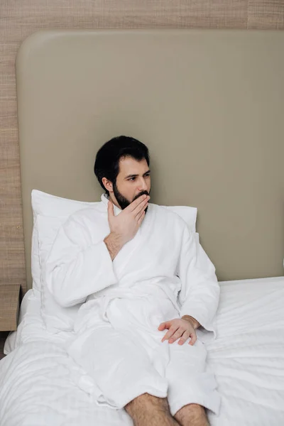 Hombre guapo en albornoz sentado en la cama en la suite del hotel y bostezando - foto de stock