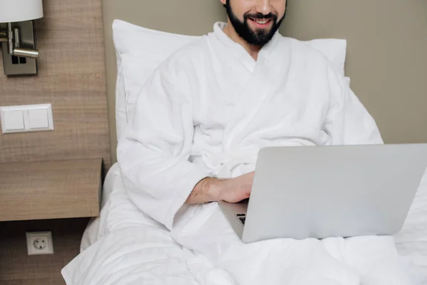 Tiro recortado de homem sorridente em roupão de banho usando laptop na cama na suíte do hotel — Fotografia de Stock