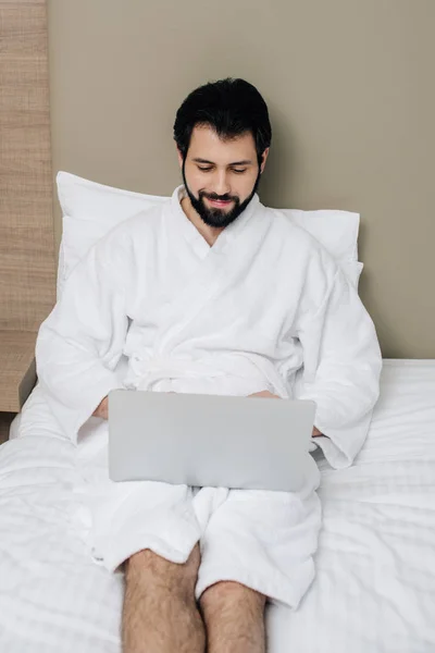 Hombre sonriente en albornoz usando el ordenador portátil en la cama en la suite del hotel - foto de stock