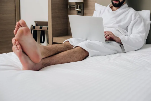 Homem bonito em roupão de banho usando laptop na cama na suíte do hotel — Fotografia de Stock