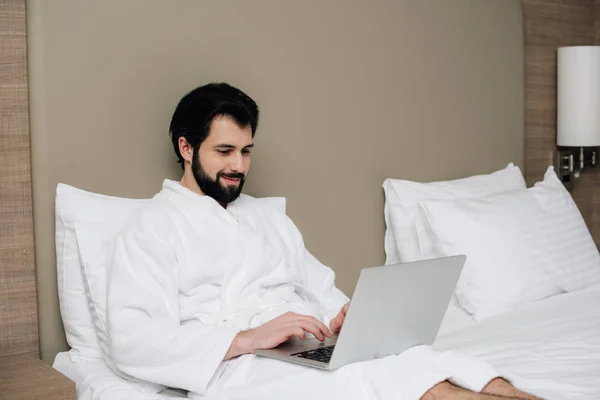 Hombre guapo en albornoz usando el ordenador portátil en la cama en la suite del hotel - foto de stock