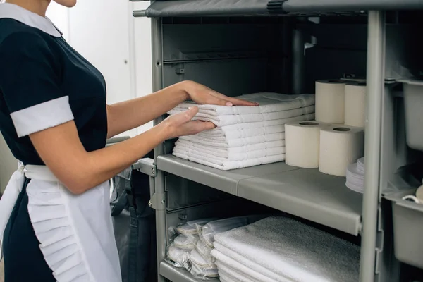 Cortado tiro de empregada doméstica em uniforme tomando toalhas de prateleira do carrinho de limpeza — Fotografia de Stock