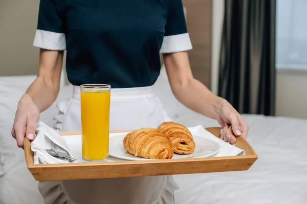 Corte tiro de empregada em uniforme segurando croissants e suco na bandeja na suíte do hotel — Fotografia de Stock