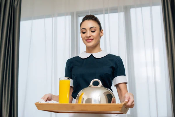 Jovem sorrindo empregada em uniforme segurando café da manhã coberto com cloche na bandeja — Fotografia de Stock