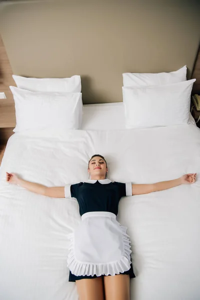 Alto ângulo vista de jovem empregada atraente em uniforme deitado na cama na suíte do hotel — Fotografia de Stock