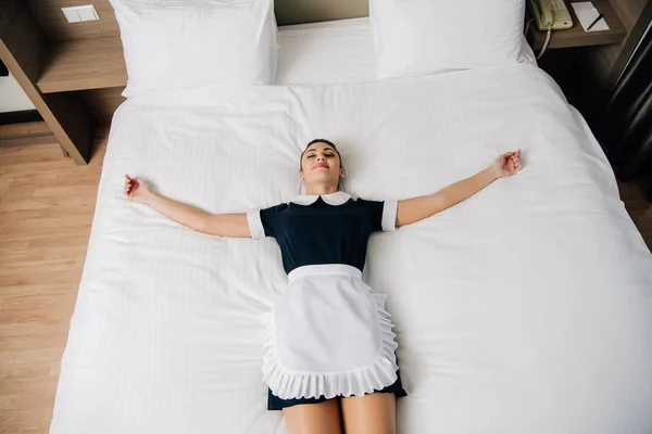 Vista ad alto angolo di giovane bella cameriera in uniforme sdraiata sul letto nella suite dell'hotel — Foto stock