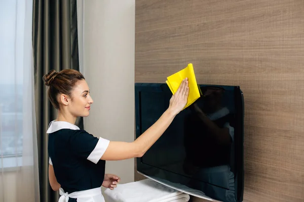 Young beautiful maid in uniform cleaning tv screen with rag — Stock Photo