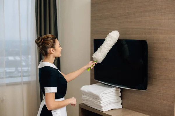 Young beautiful maid in uniform cleaning hotel suit with duster — Stock Photo