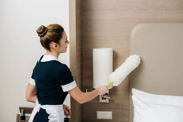 Jovem empregada doméstica atraente em uniforme terno hotel de limpeza com espanador — Fotografia de Stock