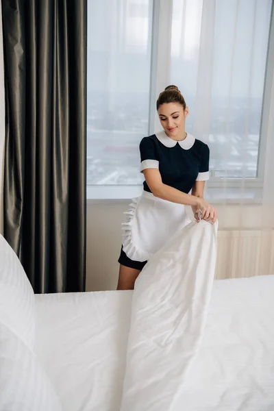 Joven hermosa criada en uniforme haciendo cama en suite de hotel - foto de stock