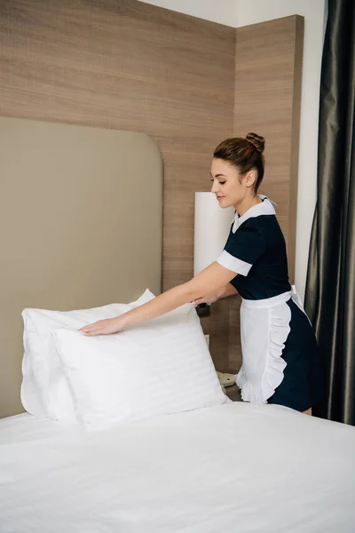 Joven feliz criada en uniforme haciendo cama en hotel suite - foto de stock