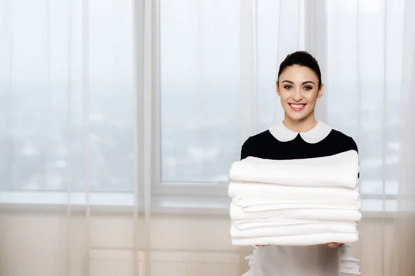Young smiling maid in uniform holding stack of clean towels and looking at camera — Stock Photo