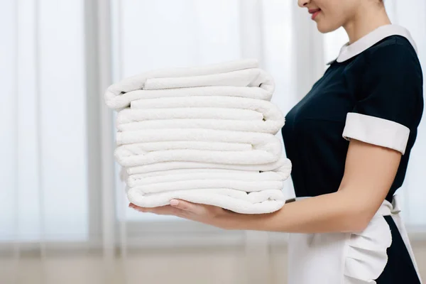 Cortado tiro de empregada em uniforme segurando pilha de toalhas limpas — Fotografia de Stock