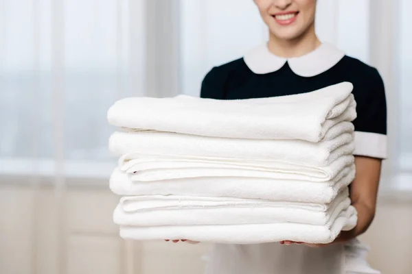 Tiro cortado de empregada sorridente em uniforme segurando pilha de toalhas limpas — Fotografia de Stock