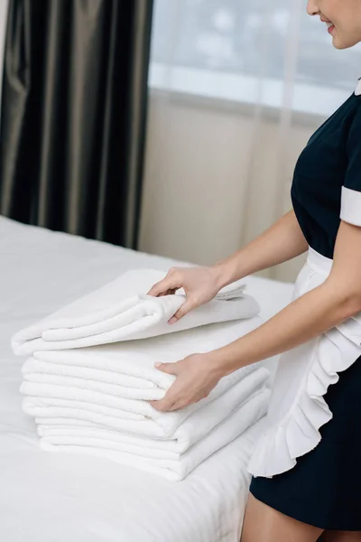 Corte tiro de empregada sorridente em uniforme com pilha de toalhas limpas na cama na suíte do hotel — Fotografia de Stock