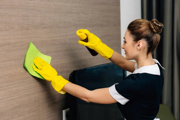 Jovem bela empregada doméstica em uniforme limpando parede de madeira com pano e aerossol limpador de móveis — Fotografia de Stock