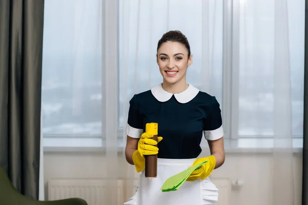 Jeune belle femme de ménage en uniforme avec chiffon et aérosol nettoyage de meubles — Photo de stock