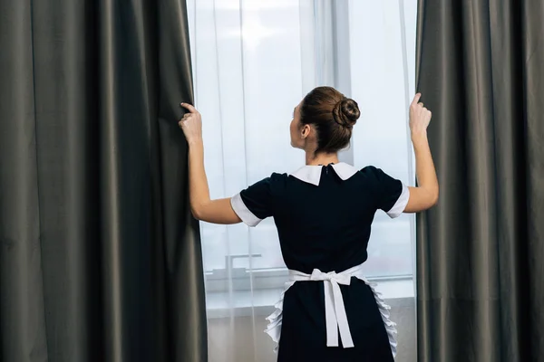 Vue arrière de jeune belle femme de ménage en uniforme rideaux de fermeture — Photo de stock