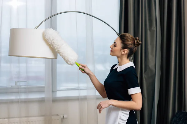 Junge glückliche Dienstmädchen in Uniform Putzlampe mit Staubwedel — Stockfoto