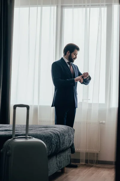 Guapo hombre de negocios en suite de hotel mirando el reloj mientras está de pie cerca de la ventana - foto de stock