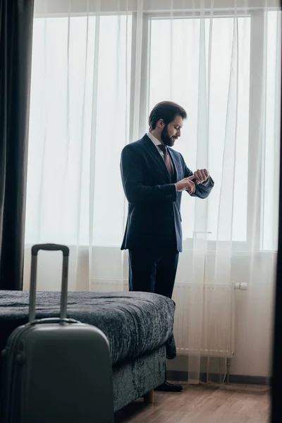 Handsome businessman in hotel suite looking at watch while standing near window — Stock Photo