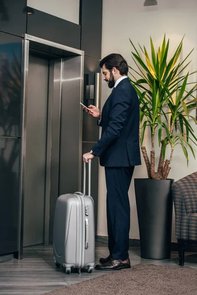 Hombre de negocios con equipaje usando el teléfono inteligente mientras espera el ascensor en el hotel - foto de stock