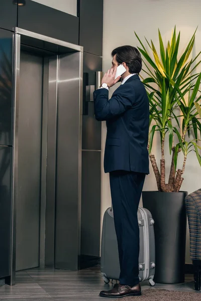 Stylish businessman with luggage talking by phone while waiting for elevator at hotel — Stock Photo