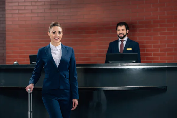 Smiling businesswoman with luggage standing in front of hotel reception counter with administrator — Stock Photo