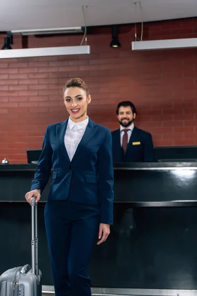 Mujer de negocios feliz con el equipaje de pie en frente del mostrador de recepción del hotel con el administrador - foto de stock