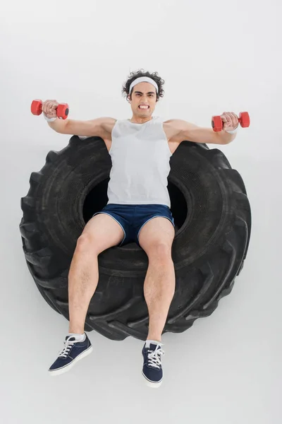 Vista de alto ângulo do atleta magro que exercita com halteres dentro do pneu da roda isolado no branco — Fotografia de Stock