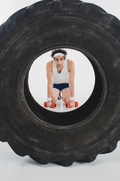Young thin sportsman sitting with dumbbells in hands with tire on foreground — Stock Photo
