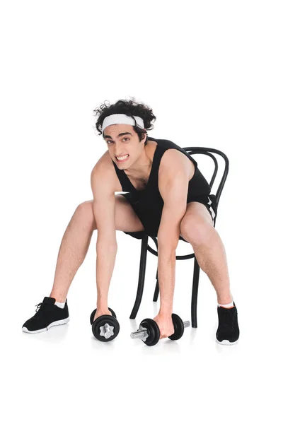 Skinny sportsman trying to raising dumbbells while sitting on chair isolated on white — Stock Photo