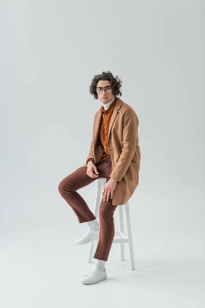 Jeune homme élégant dans des lunettes assis sur une chaise isolée sur gris — Photo de stock