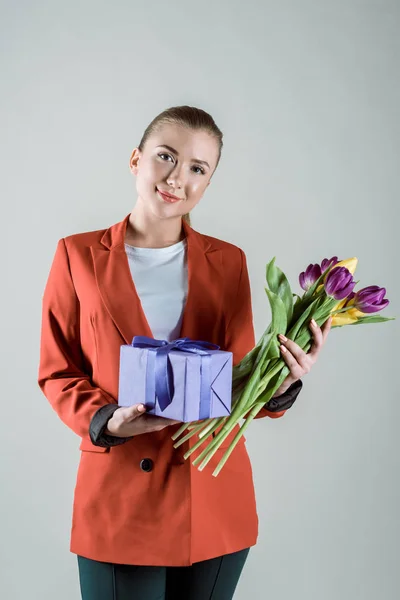 Happy woman holding gift box and flower bouquet isolated on grey — Stock Photo