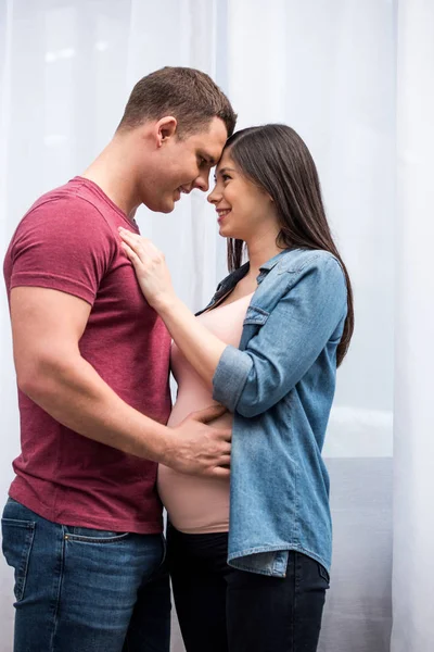 Vista laterale della bella felice giovane coppia incinta che si abbraccia e sorride a casa — Foto stock