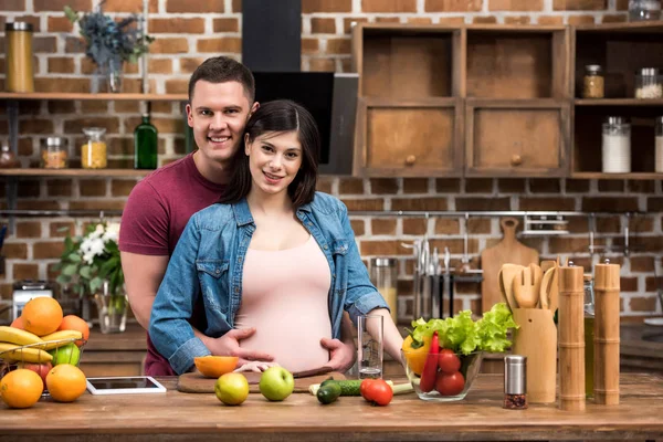 Beautiful happy young pregnant couple hugging and smiling at camera in kitchen — Stock Photo