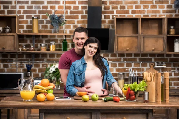 Bella giovane coppia incinta abbracciare e sorridere alla macchina fotografica mentre si cucina insieme — Foto stock