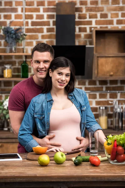 Heureux jeune couple enceinte étreignant et souriant à la caméra tout en cuisinant ensemble — Photo de stock