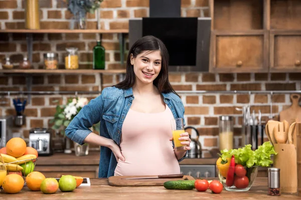 Joven embarazada sosteniendo un vaso de jugo fresco y sonriendo a la cámara en la cocina - foto de stock