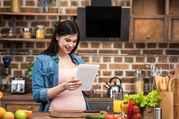 Sourire jeune femme enceinte en utilisant une tablette numérique tout en cuisinant à la cuisine — Photo de stock