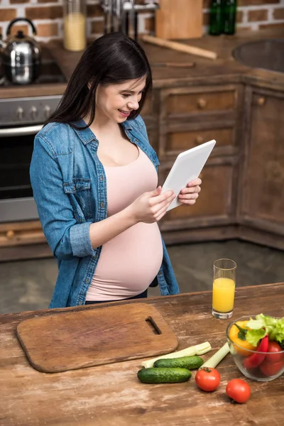 Smiling young pregnant woman using digital tablet while cooking at kitchen — Stock Photo