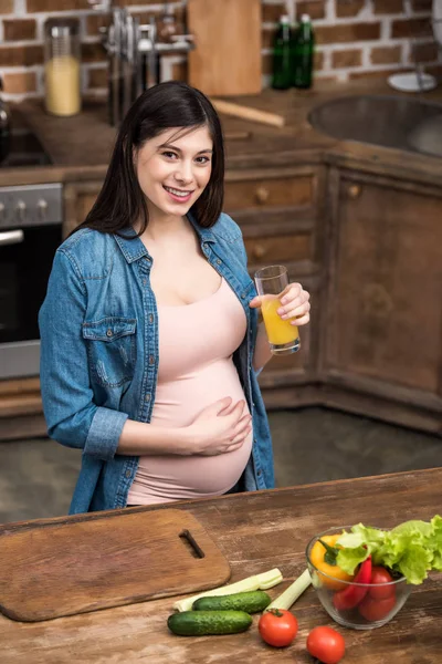 Hermosa joven embarazada sosteniendo un vaso de jugo fresco y sonriendo a la cámara - foto de stock
