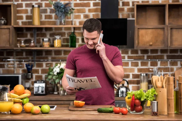 Jovem feliz falando por smartphone e lendo jornal de viagem na cozinha — Fotografia de Stock