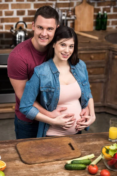 Heureux jeune couple enceinte étreignant et souriant à la caméra à la cuisine — Photo de stock