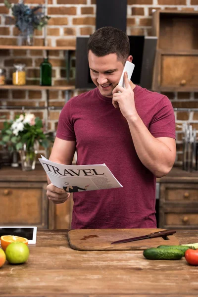 Joven sonriente leyendo el periódico de viaje y hablando por teléfono inteligente - foto de stock