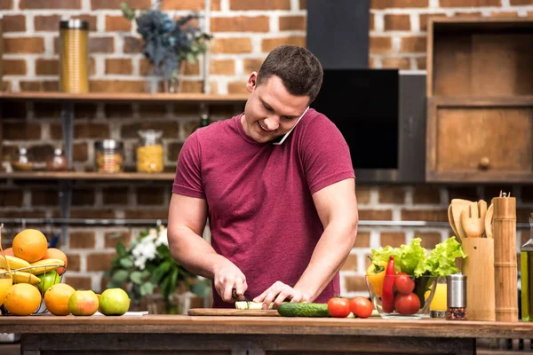 Smiling young man talking by smartphone and cooking salad in kitchen — Stock Photo