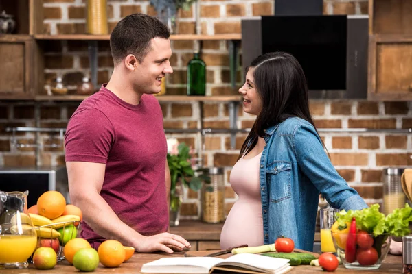 Seitenansicht eines glücklichen jungen schwangeren Paares, das einander beim gemeinsamen Kochen in der Küche lächelt — Stockfoto