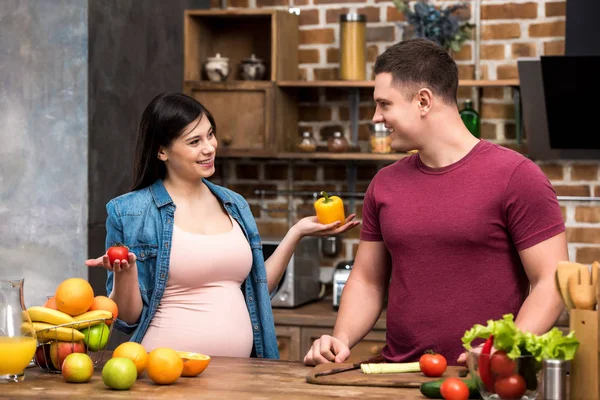 Щаслива молода вагітна пара посміхається один одному, готуючи разом на кухні — стокове фото